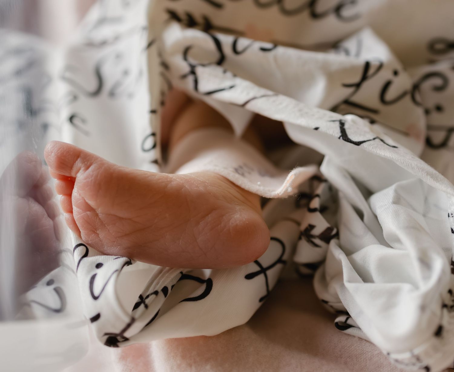 Close-up of a newborn baby's tiny foot peeking out from a white swaddle blanket with cursive text print.