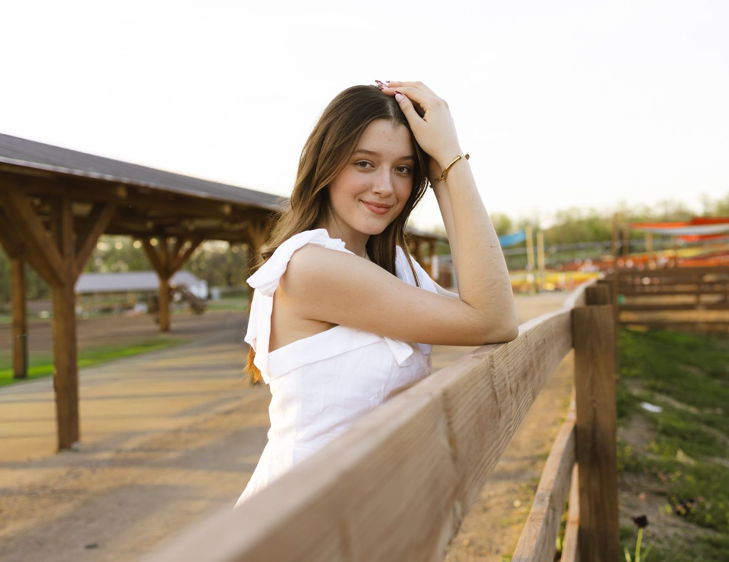 Young woman in white dress leaning on wooden fence at golden hour, smiling outdoors at a rustic farm venue.