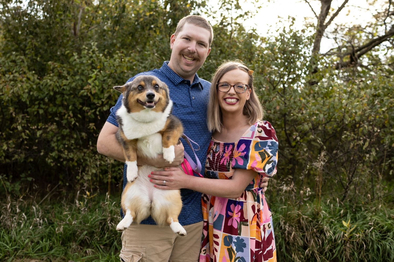 Smiling couple holding an adorable Corgi dog outdoors with lush green trees in the background.