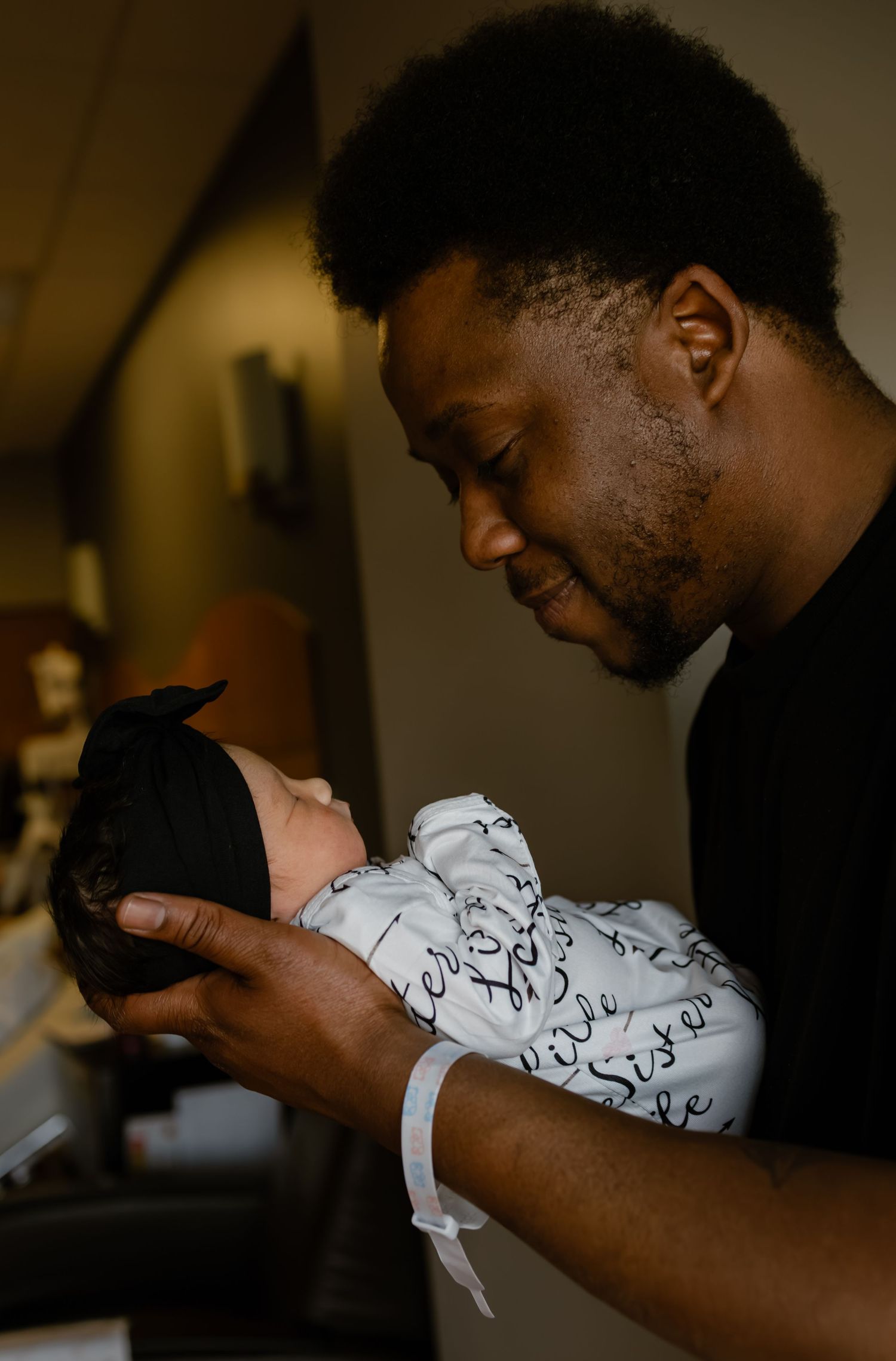 A father tenderly holds his swaddled newborn baby in a hospital room, smiling down with joy.