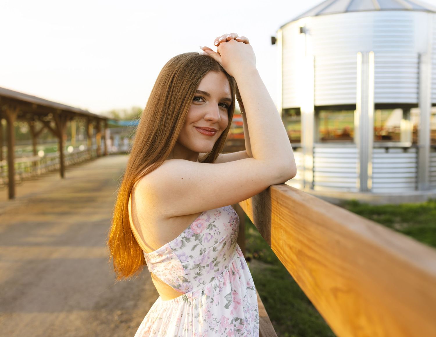 Young woman in floral sundress posing by wooden fence at golden hour with grain silos in background.
