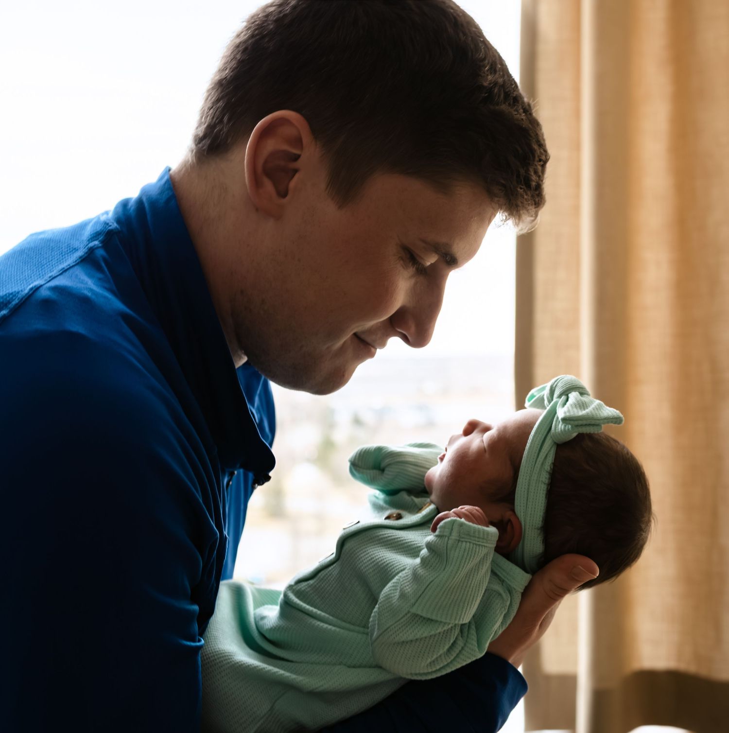 New father in blue jacket tenderly holding his newborn baby by a hospital window in soft light for their Fresh 48 Session in Omaha, NE.