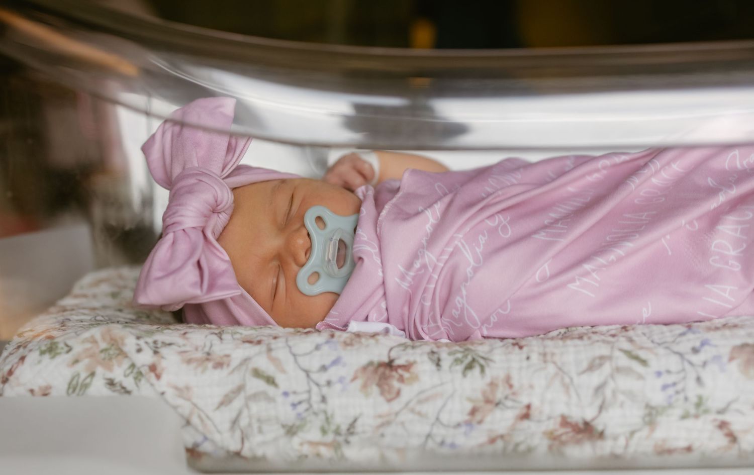 Newborn baby girl in pink swaddle and bow headband sleeping peacefully in hospital bassinet with pacifier.