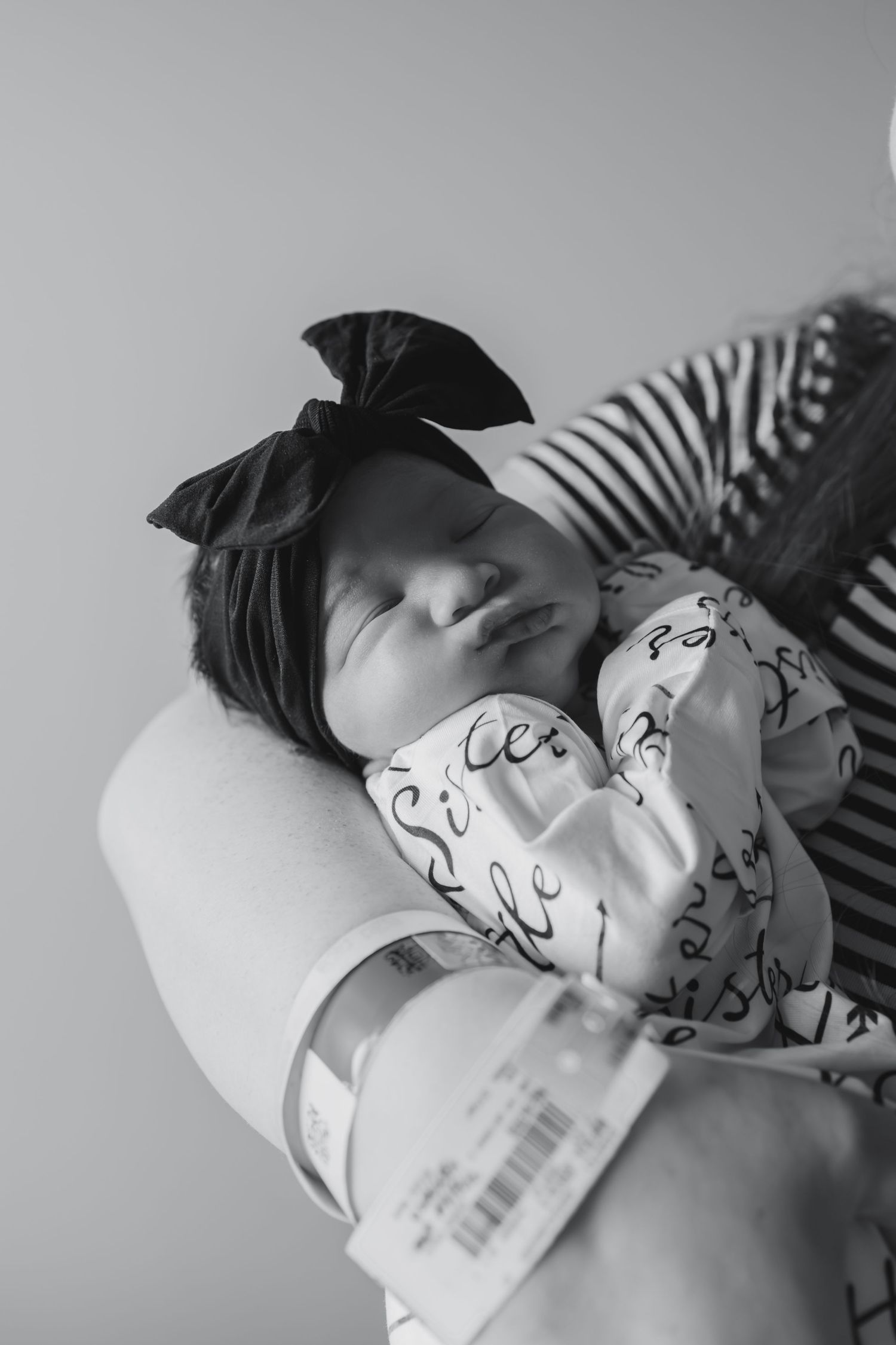 Black and white photo of a sleeping newborn baby wearing a bow headband and printed onesie, cradled in loving arms.