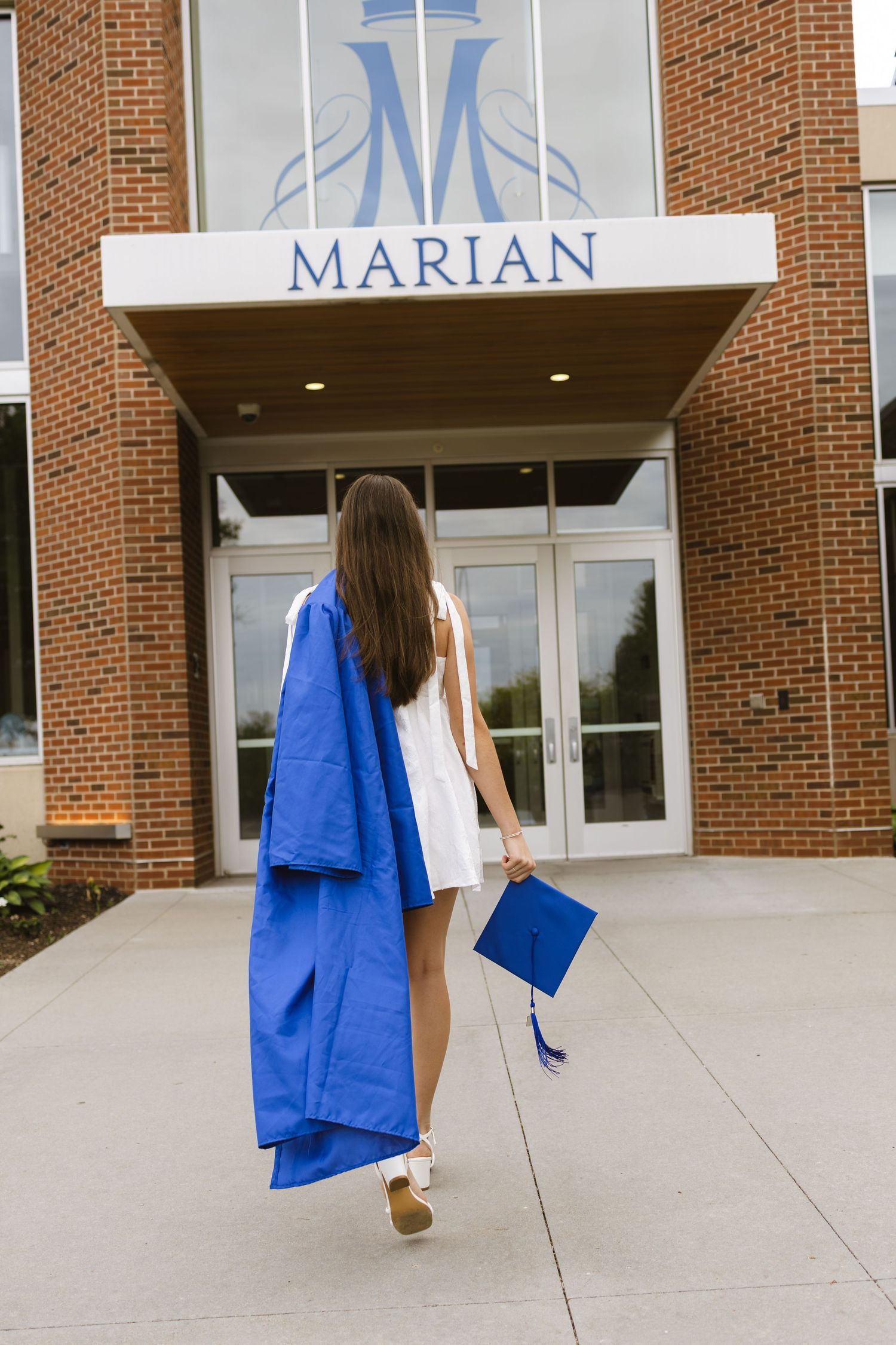 A graduate in blue cap and gown walks toward Marian University entrance holding her graduation cap.