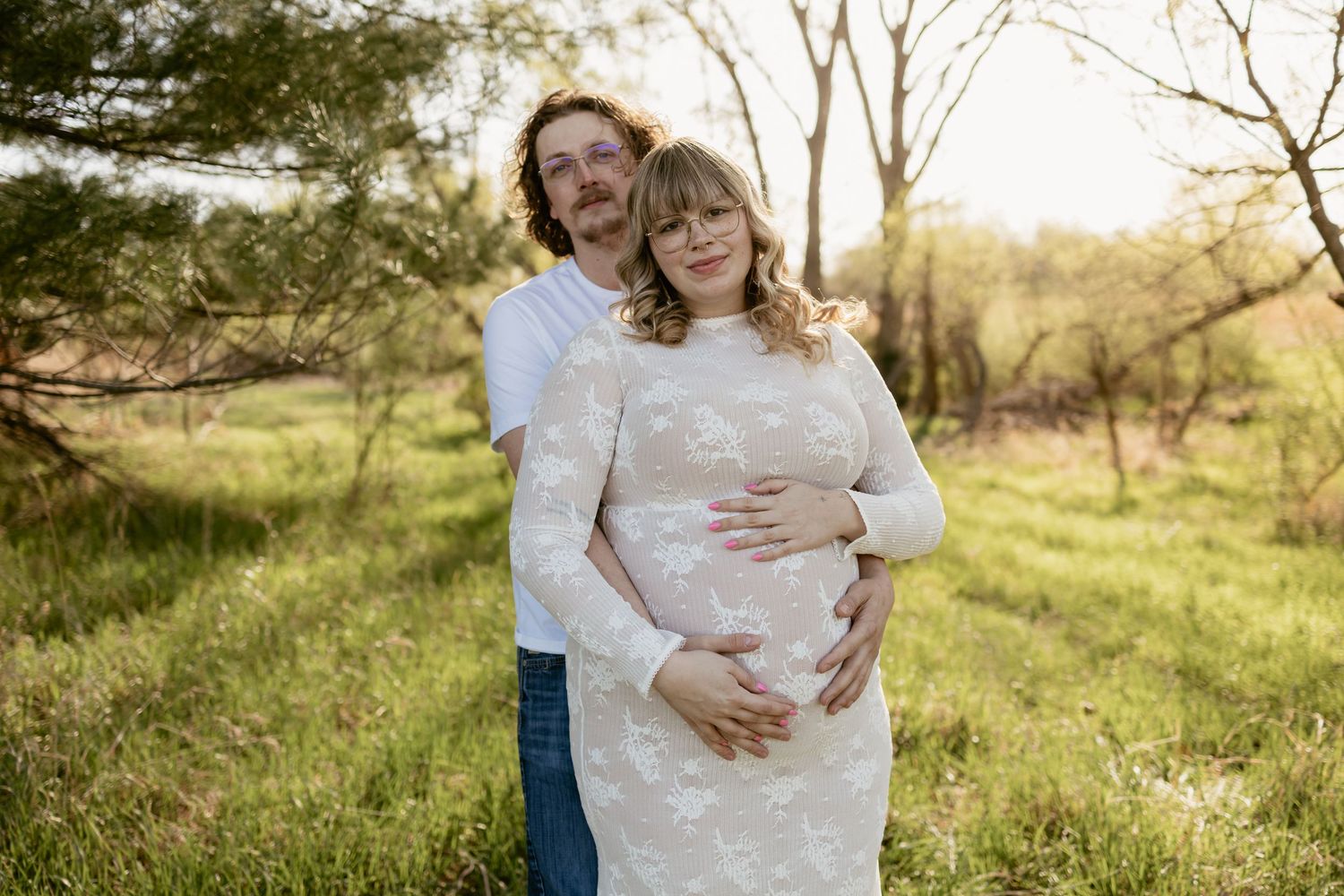 Couple posing in a sunlit field, woman in white lace dress cradling baby bump, man embracing her from behind.