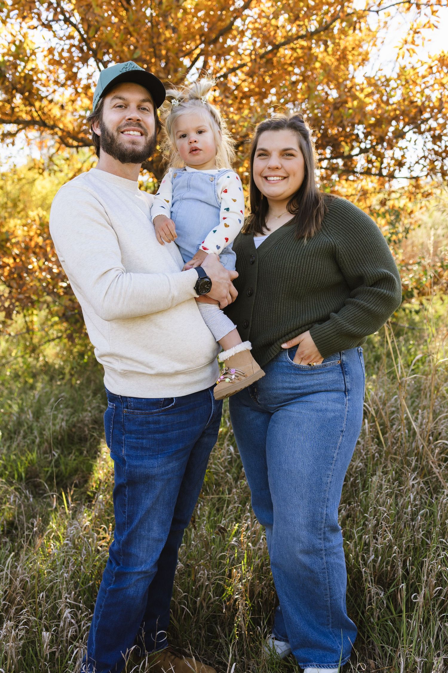 Young family of three poses outdoors in fall foliage, parents holding toddler in blue outfit, wearing jeans and casual sweaters.