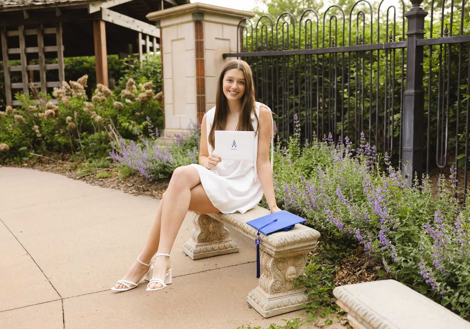 Young woman in white dress holding graduation cap sits on stone bench surrounded by purple flowers.