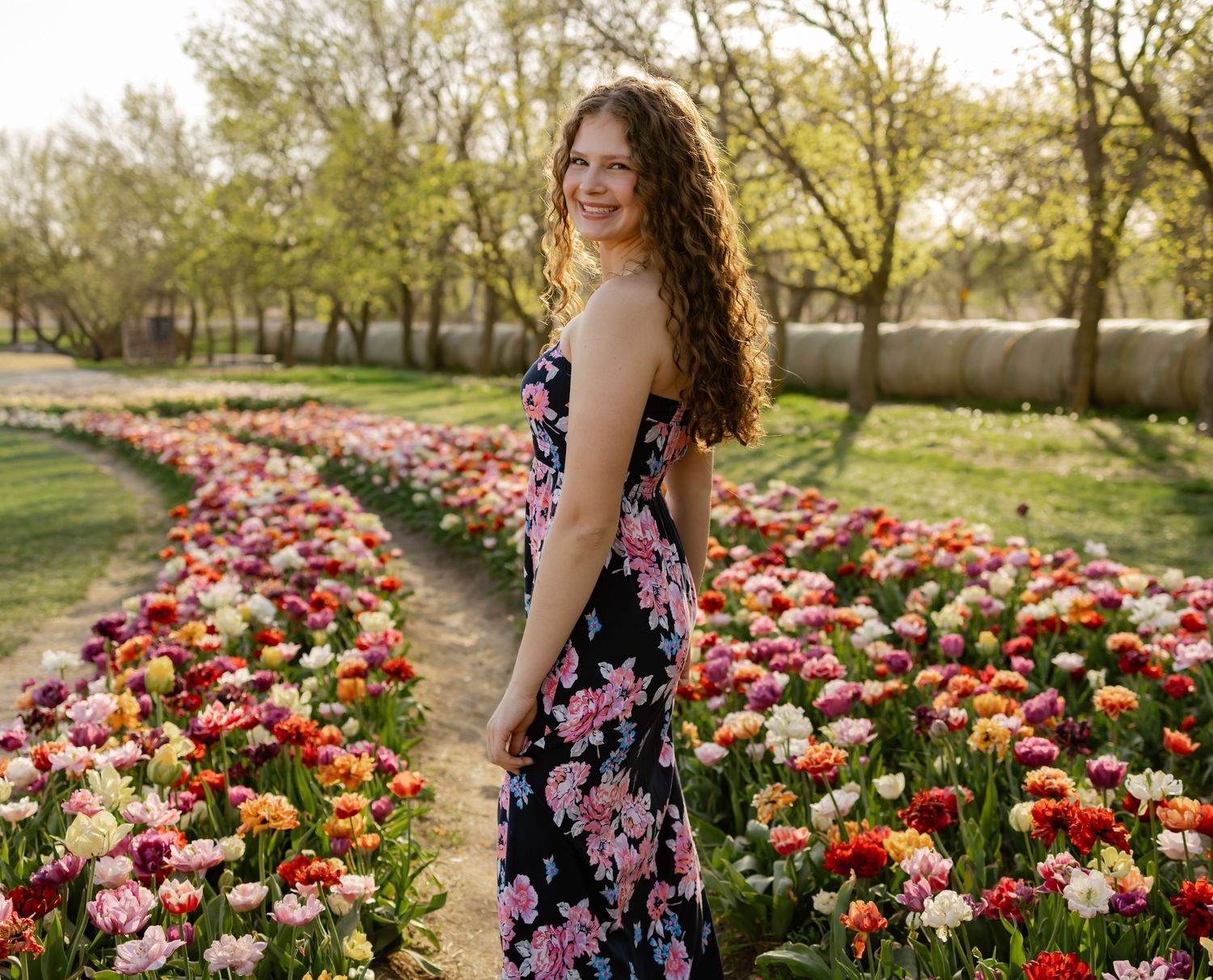 Young woman in floral dress smiling in a vibrant tulip field with colorful blooms and green trees in spring.