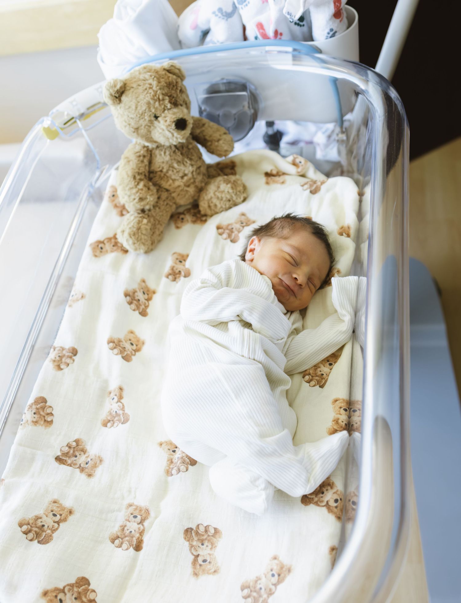 Newborn baby sleeping peacefully in a hospital bassinet with a teddy bear blanket and stuffed bear toy.