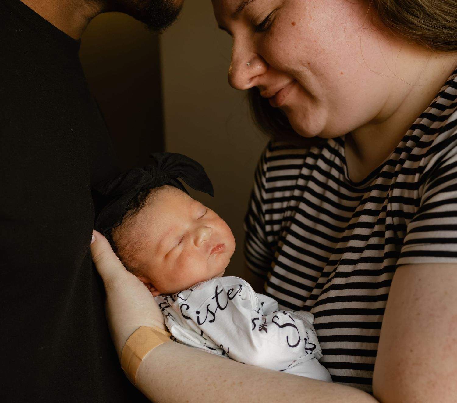 Parents lovingly cradling their sleeping newborn baby wrapped in a white swaddle blanket.