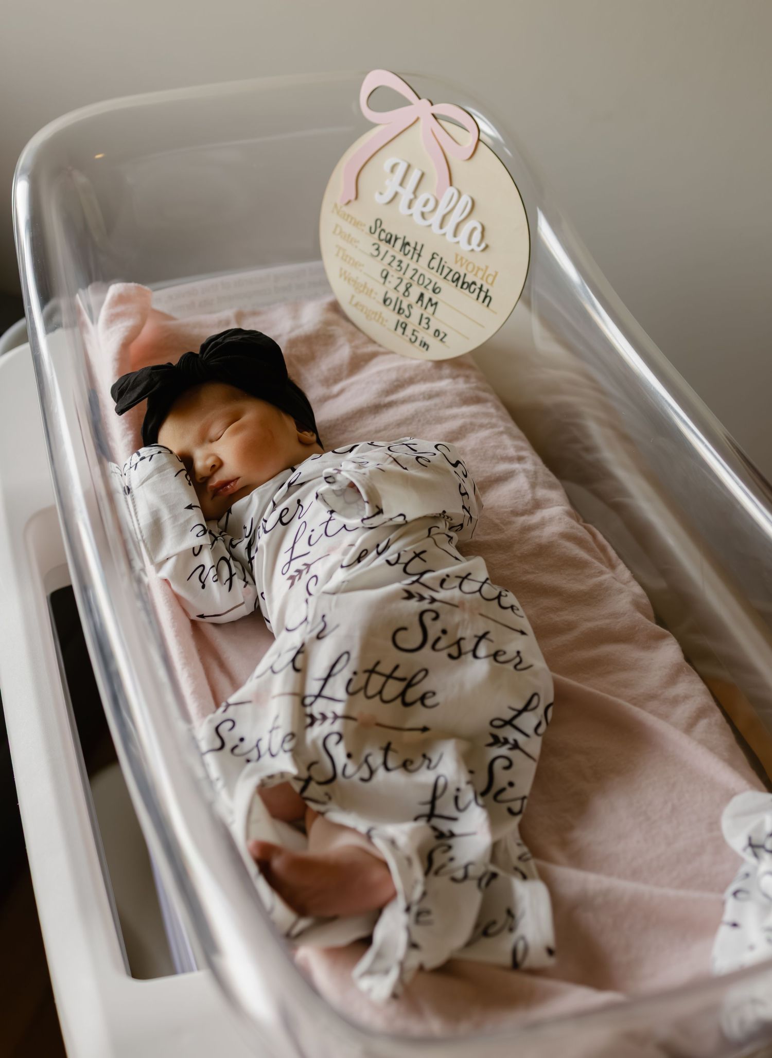 Newborn baby girl sleeping in hospital bassinet wrapped in Little Sister swaddle with pink birth announcement sign.