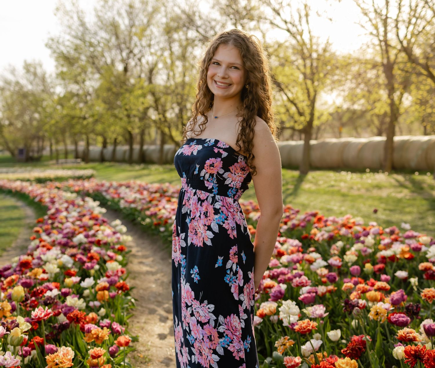 Young woman in floral maxi dress smiling in a colorful tulip garden with trees in the background.