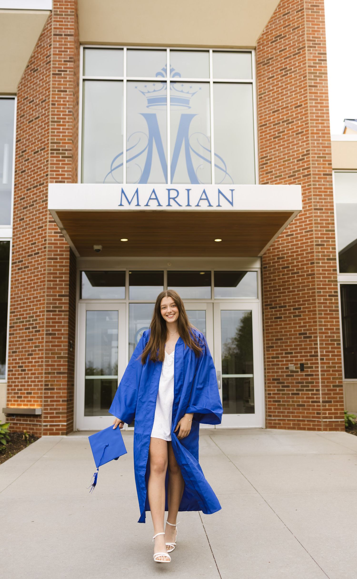 A smiling graduate in a blue cap and gown poses in front of Marian school entrance holding her graduation cap.