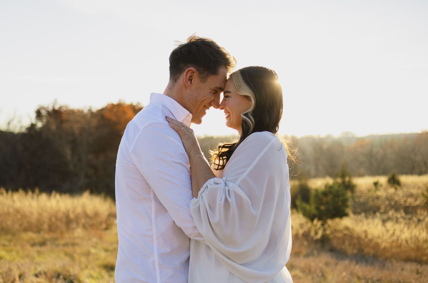 A romantic couple in Nebraska stands forehead to forehead at golden hour in an open field with fall foliage for their photos.