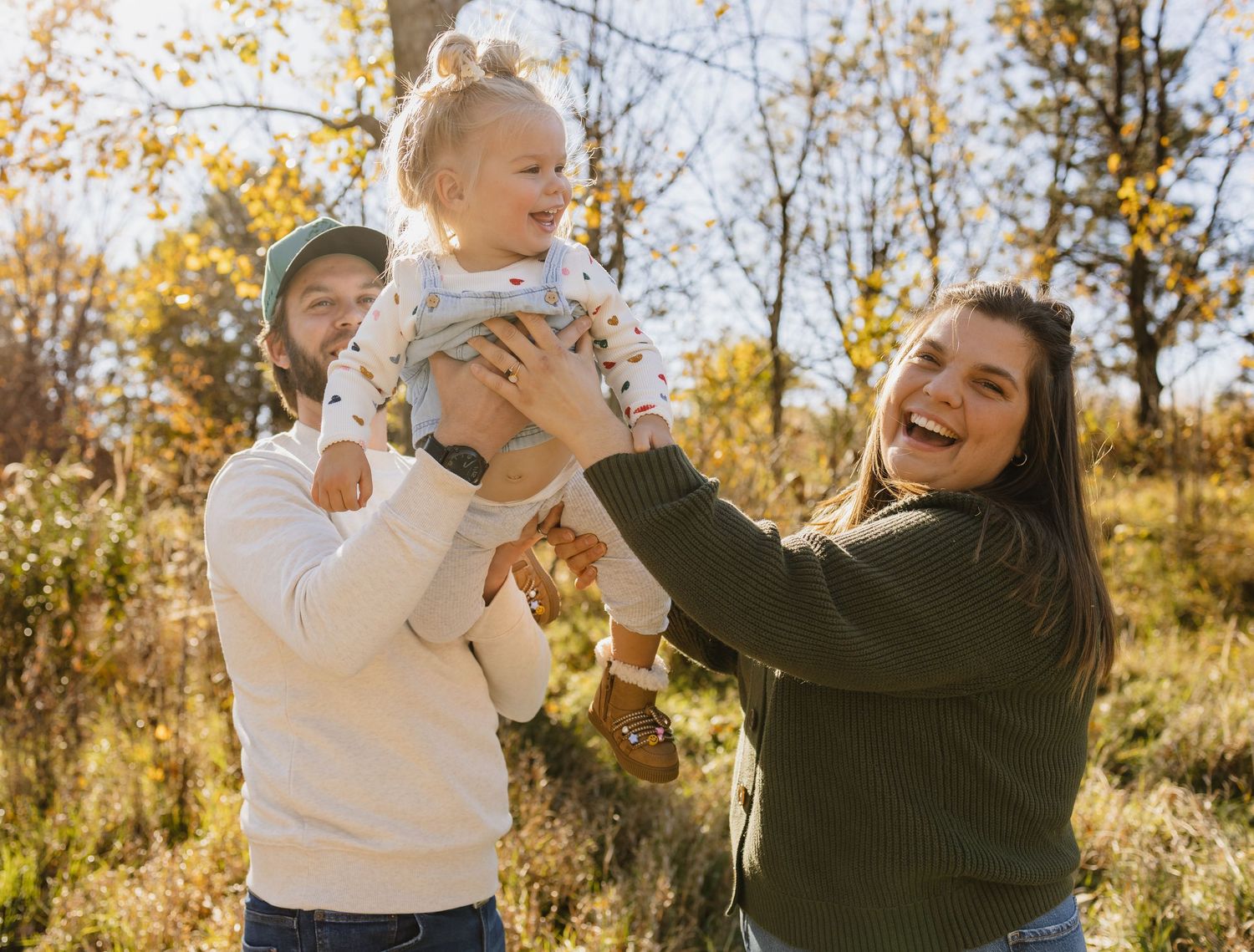 A joyful family of three enjoys a sunny autumn day outdoors, with parents lifting their laughing toddler in the air.