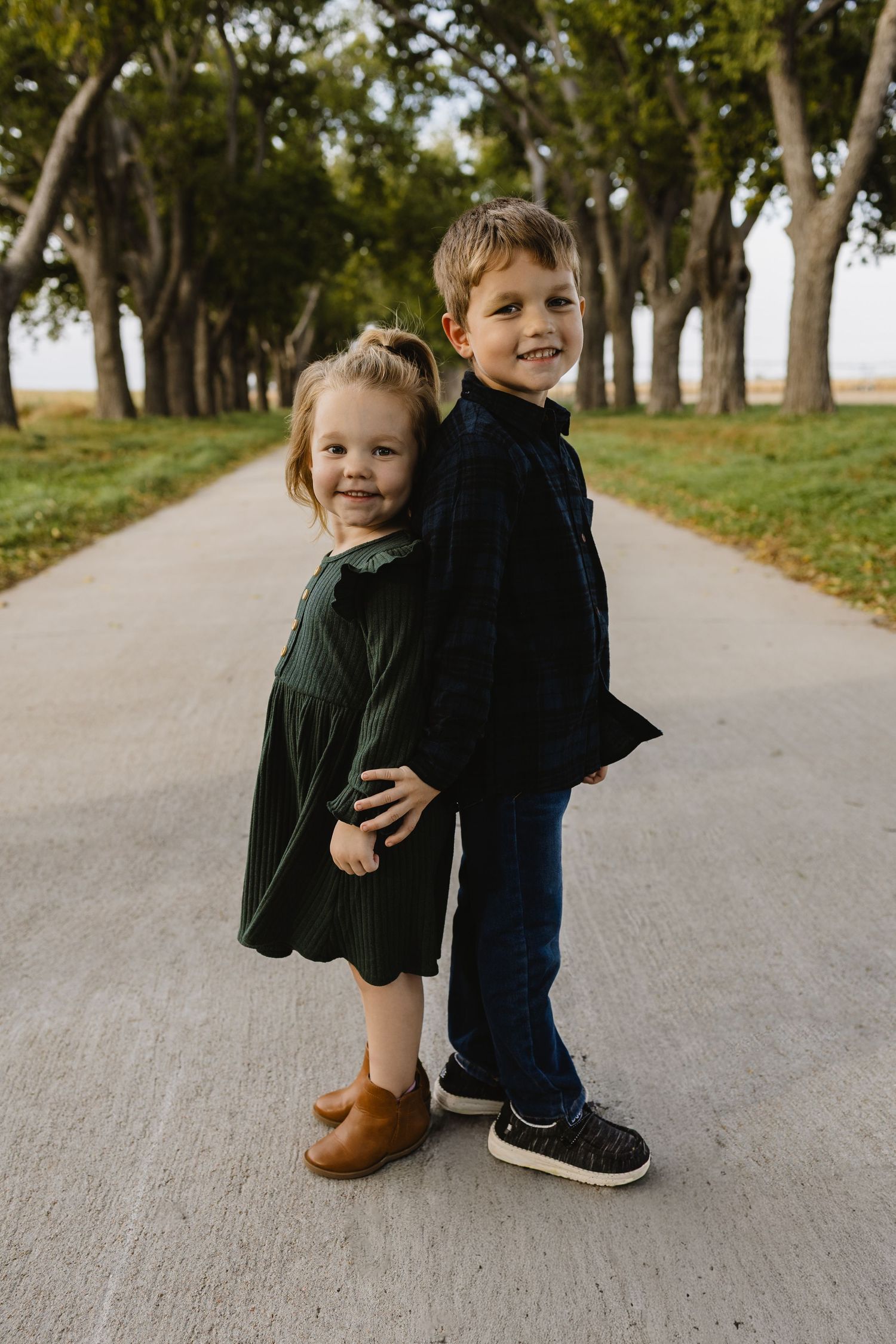 Two young siblings pose back-to-back on a tree-lined path, girl in green dress and boy in dark outfit smiling.
