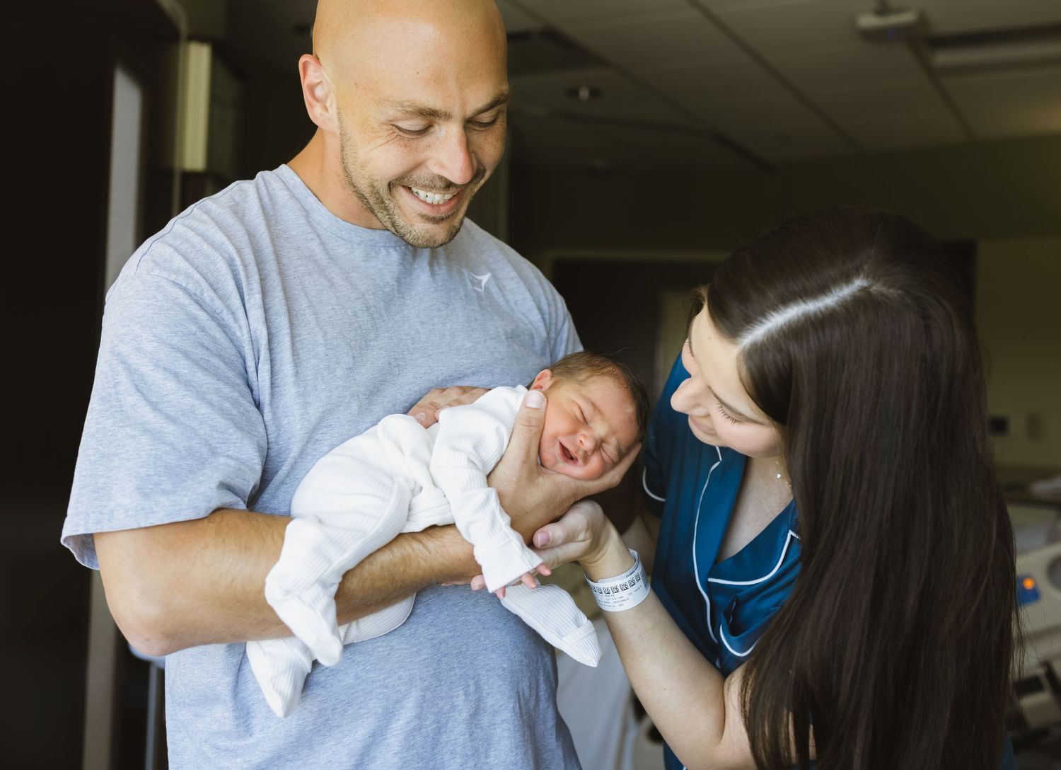 A bald man and dark-haired woman in scrubs smile while holding a swaddled newborn baby in a hospital room.