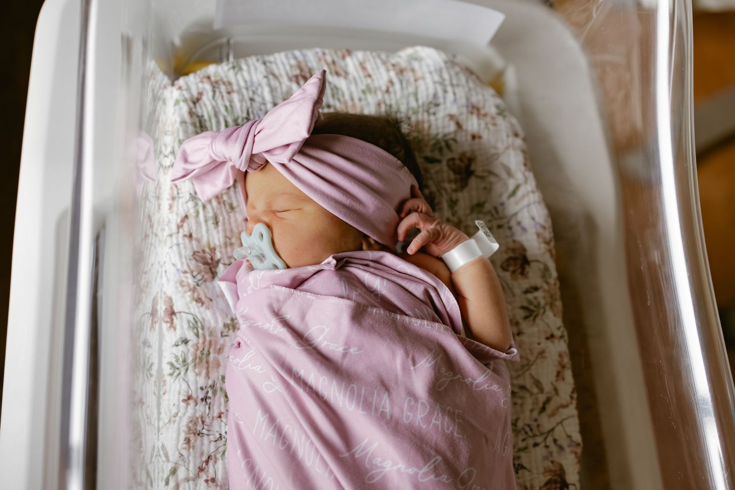 Newborn baby swaddled in pink blanket with bow headband sleeping peacefully in hospital bassinet.