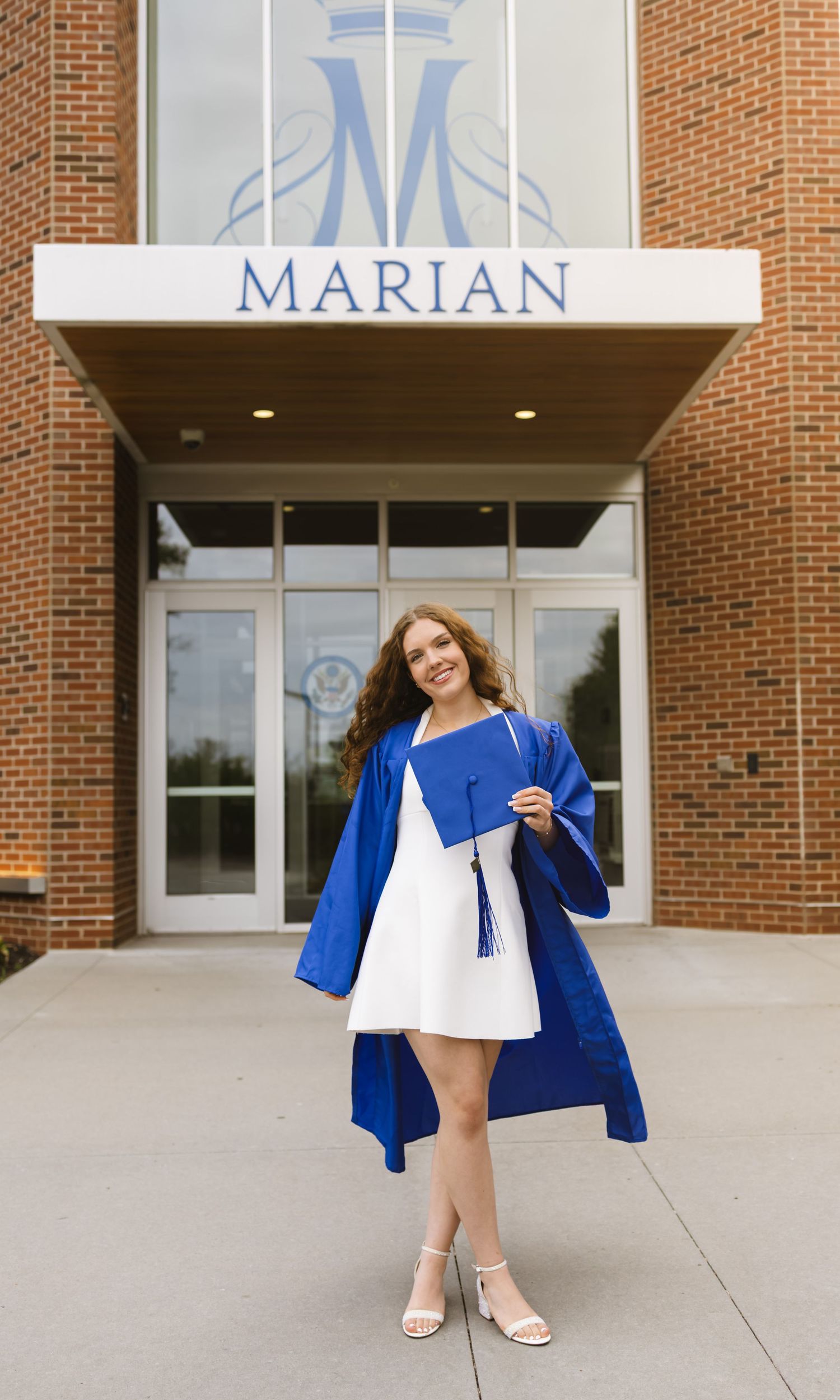 A smiling graduate in a blue cap and gown holds her diploma in front of Marian school entrance.