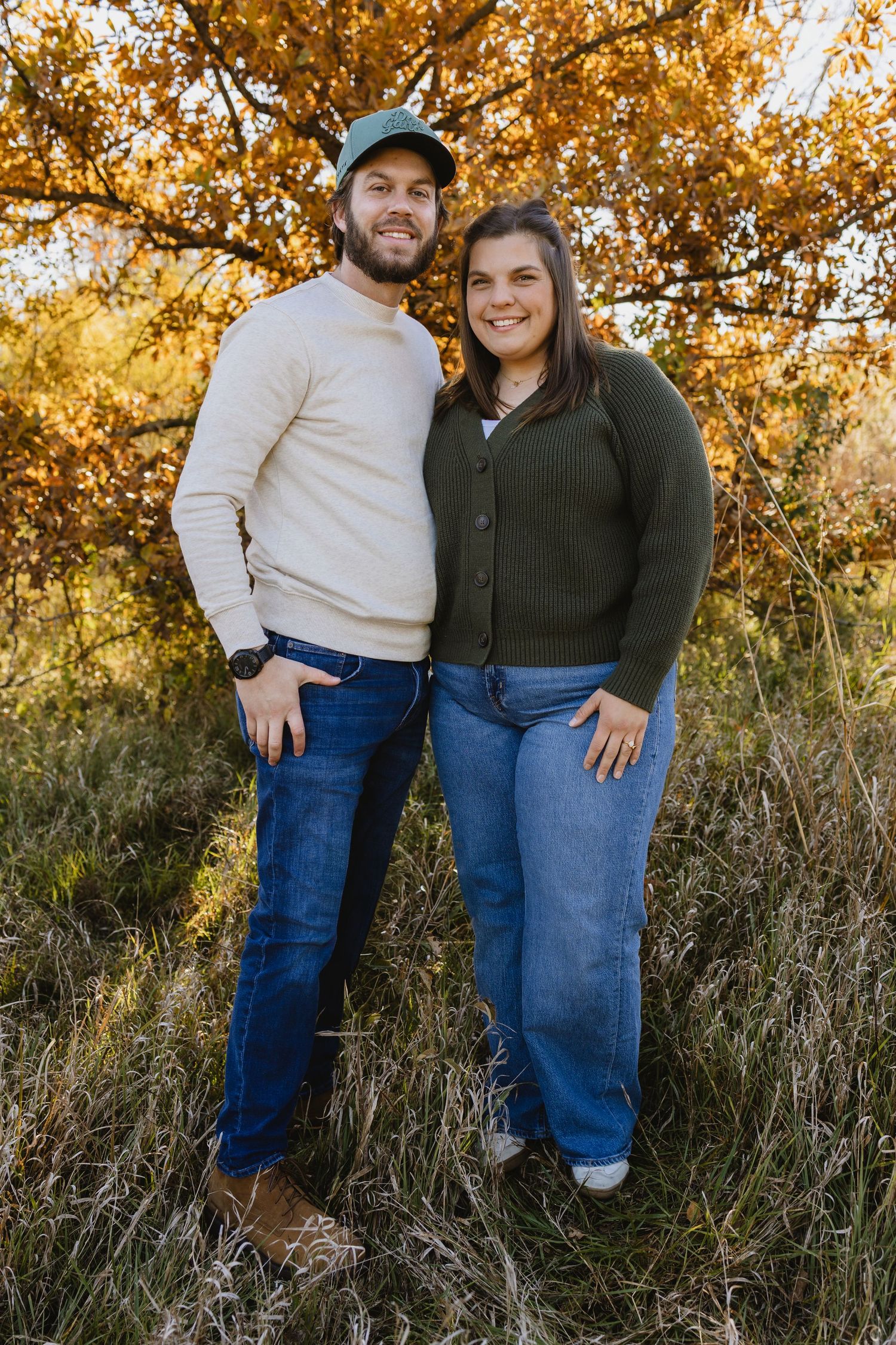 Couple posing outdoors in fall foliage, wearing casual sweaters and jeans, smiling warmly in golden autumn light.