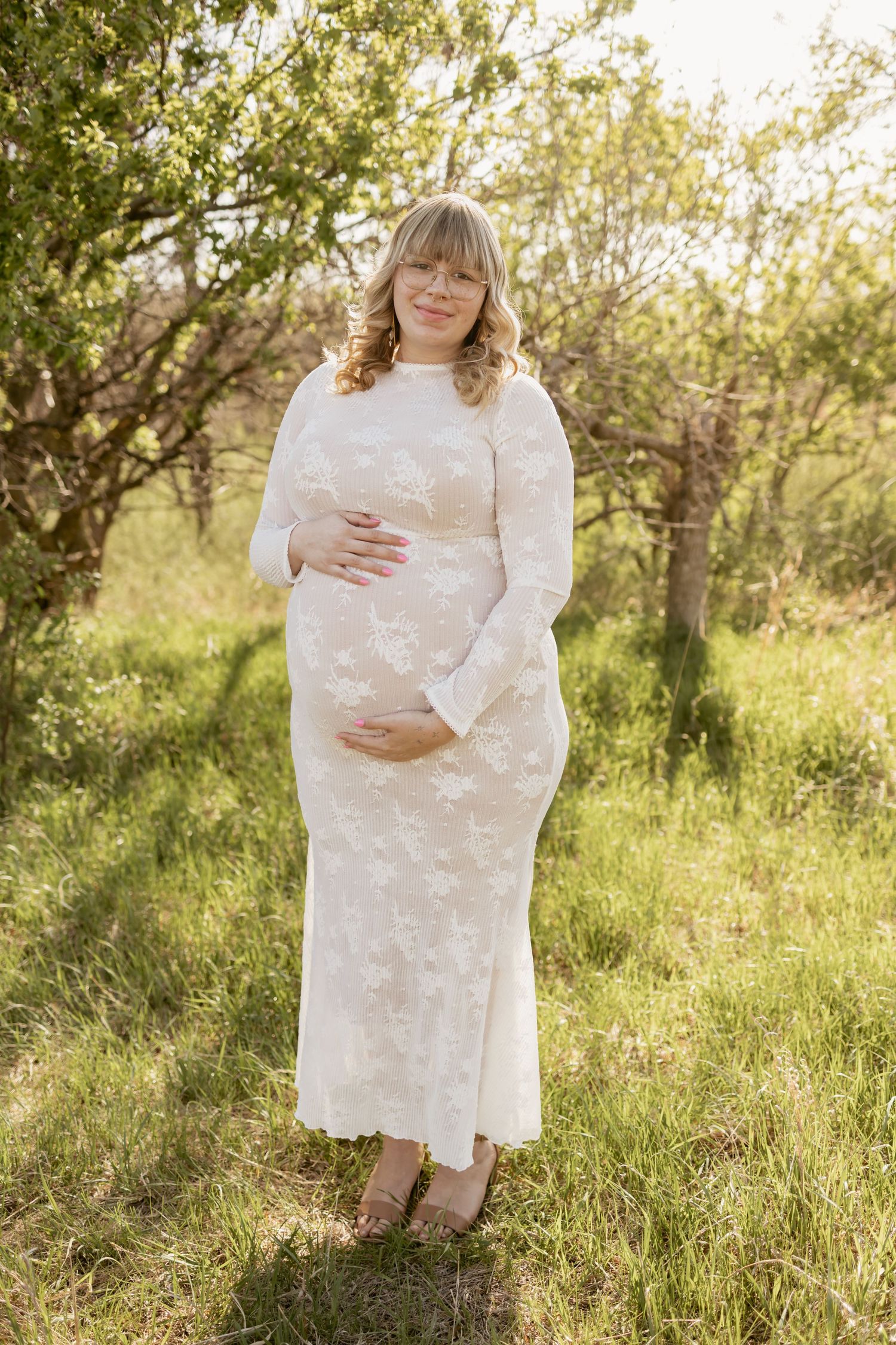 Pregnant woman in white lace dress holding belly outdoors in sunny garden.