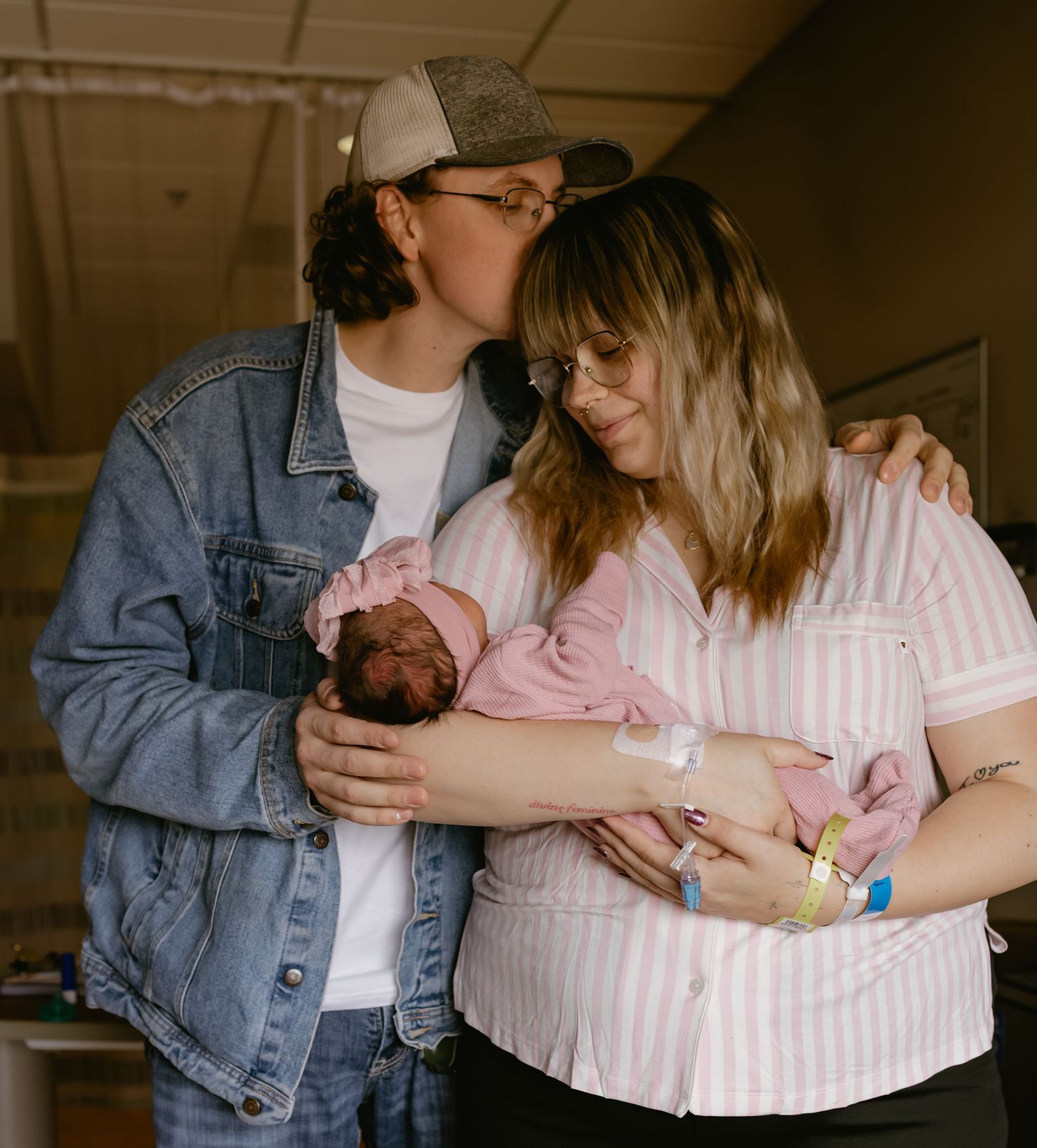 Two parents share a tender moment holding their newborn baby in a hospital room, mom in pink shirt.