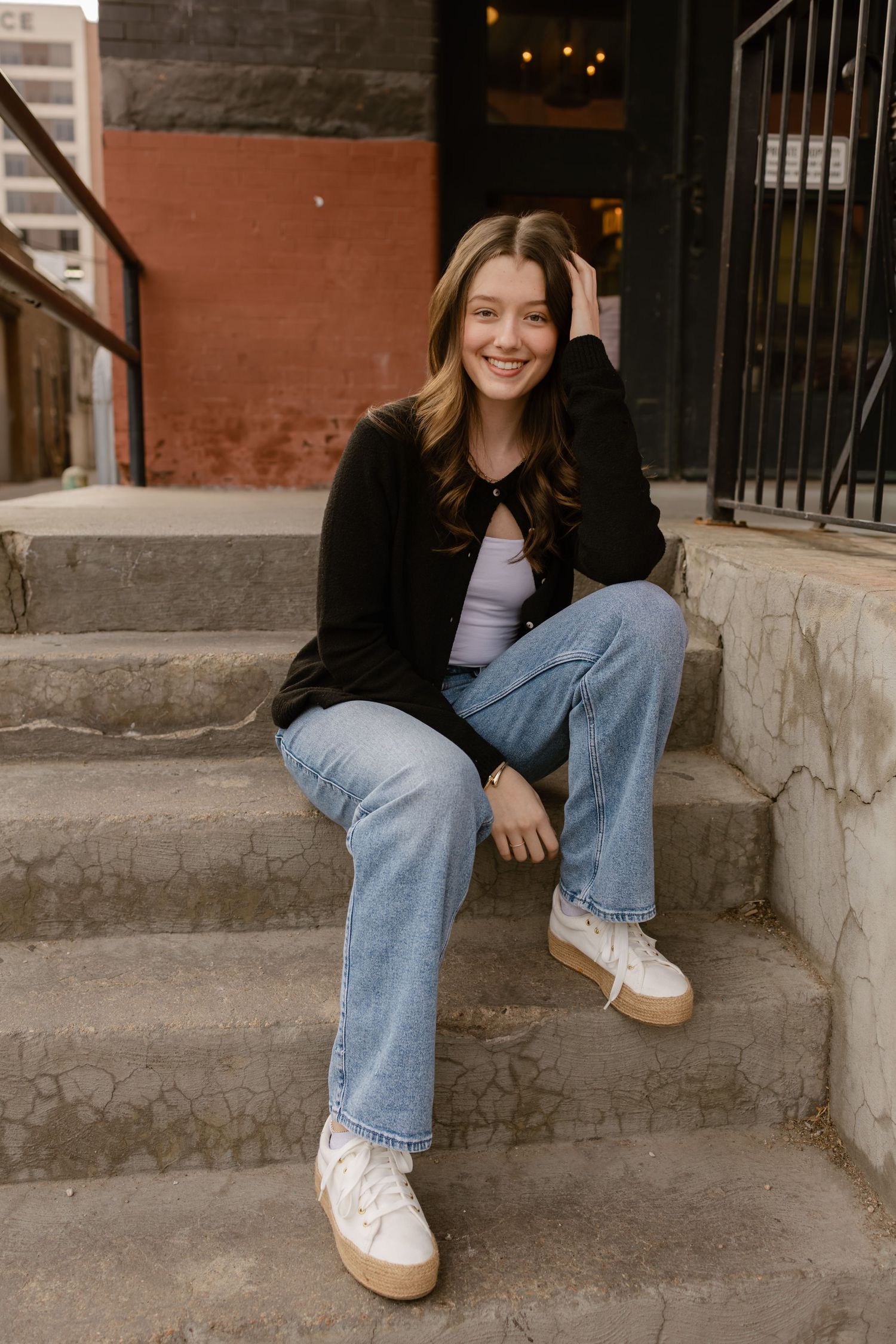 Young woman smiling on urban steps, wearing black blazer, light jeans, and white sneakers.