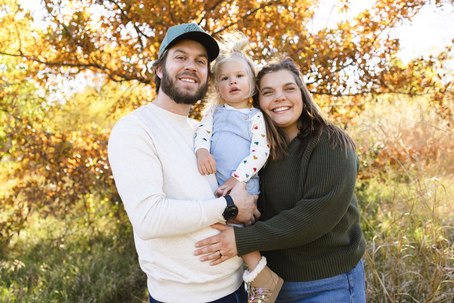 Young couple holding baby outdoors surrounded by vibrant golden autumn foliage.