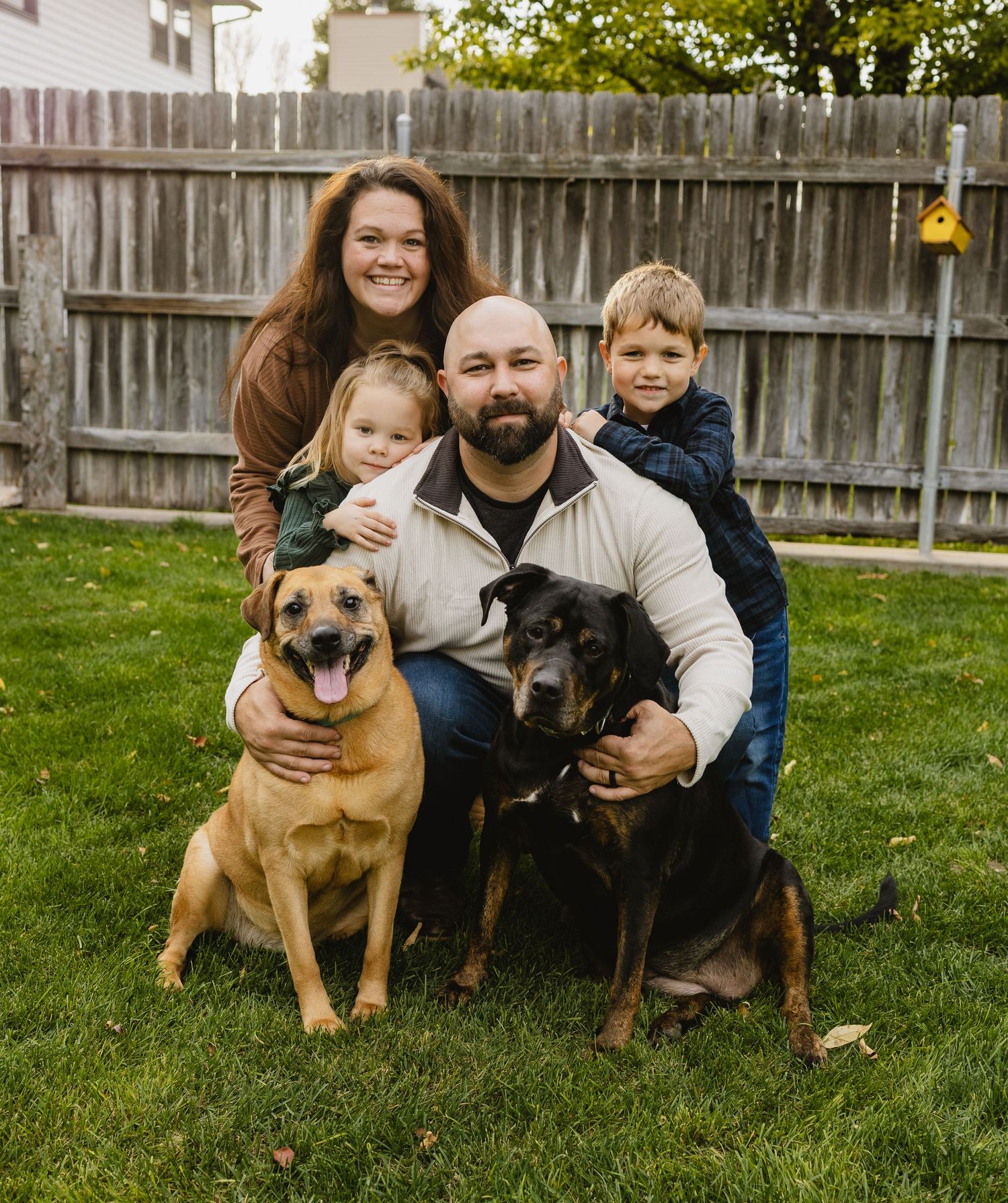 Family of four poses outdoors with two dogs on green grass, wooden fence backdrop, casual fall attire.