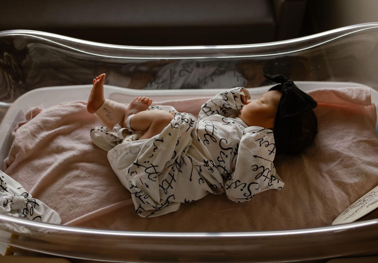 Newborn baby lying in hospital bassinet wrapped in white patterned swaddle blanket with dark hair visible.