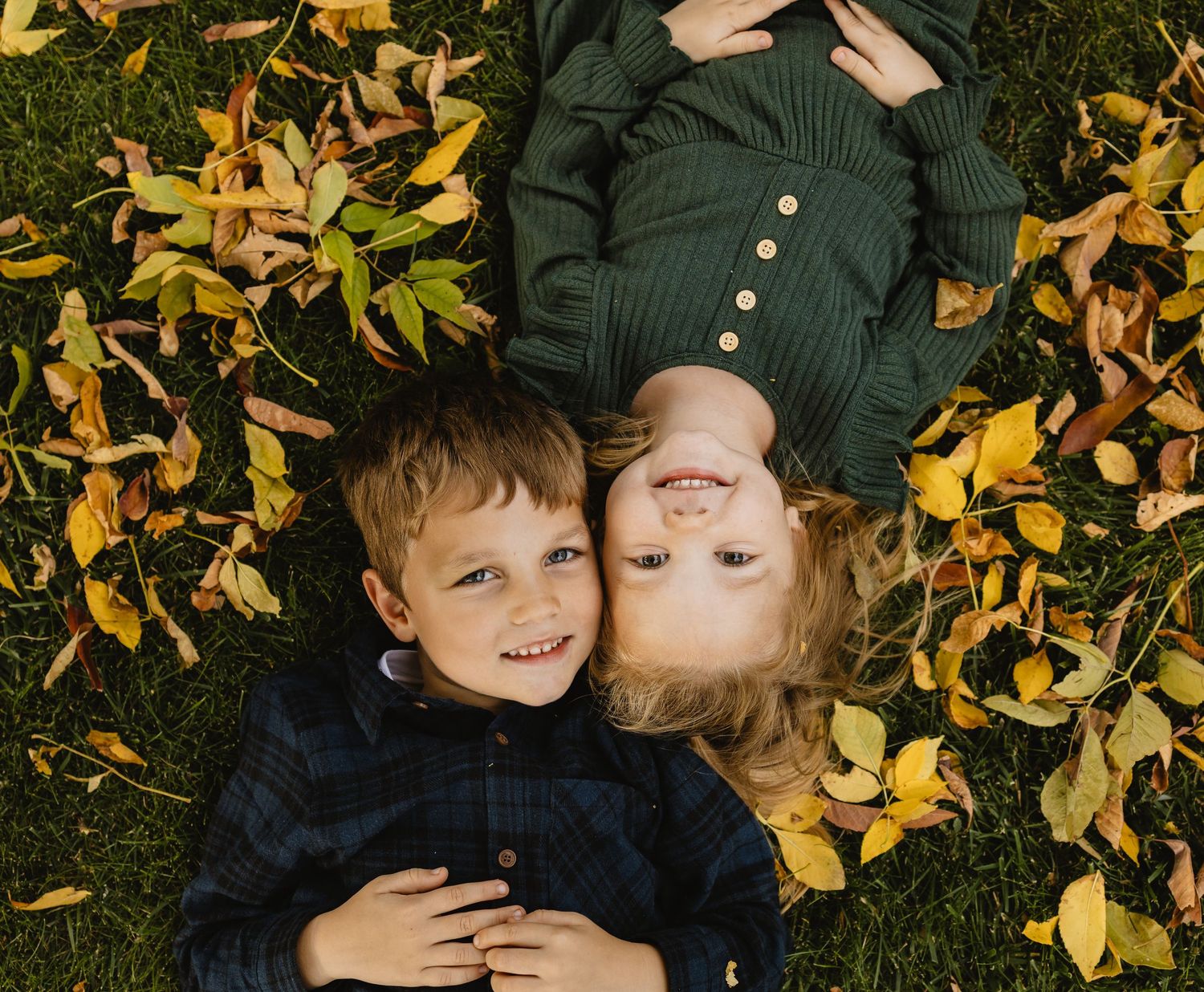 Two children lying in autumn leaves on green grass, smiling together in a cozy fall outdoor setting.