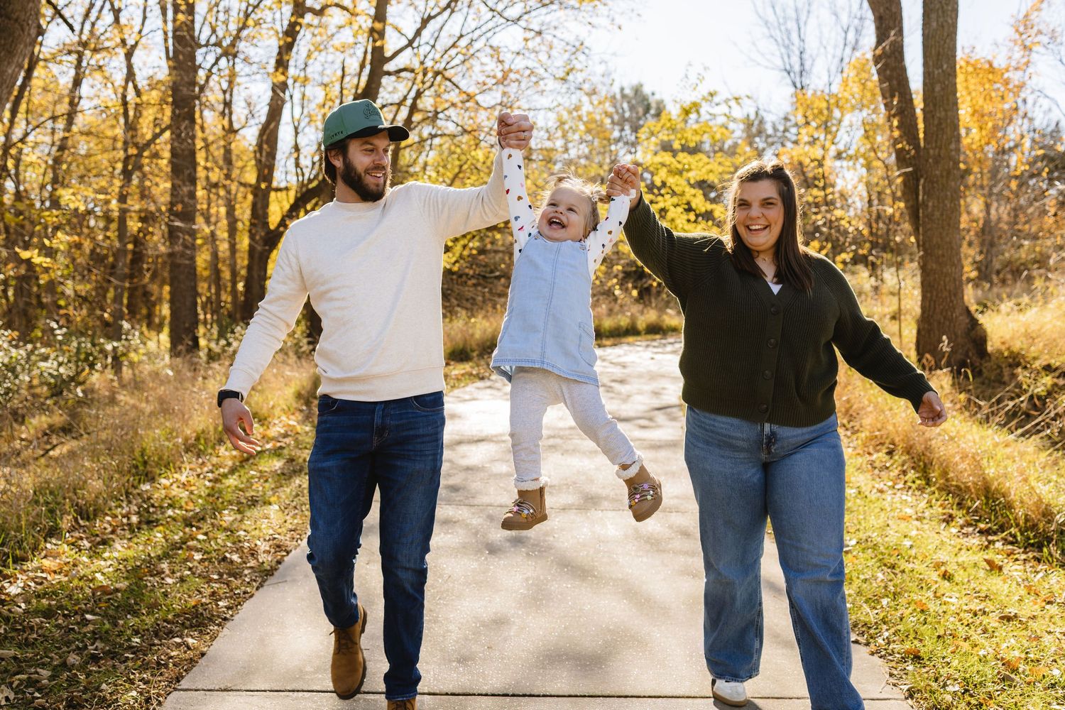 Family swinging toddler on autumn park path surrounded by golden trees on a sunny fall day.