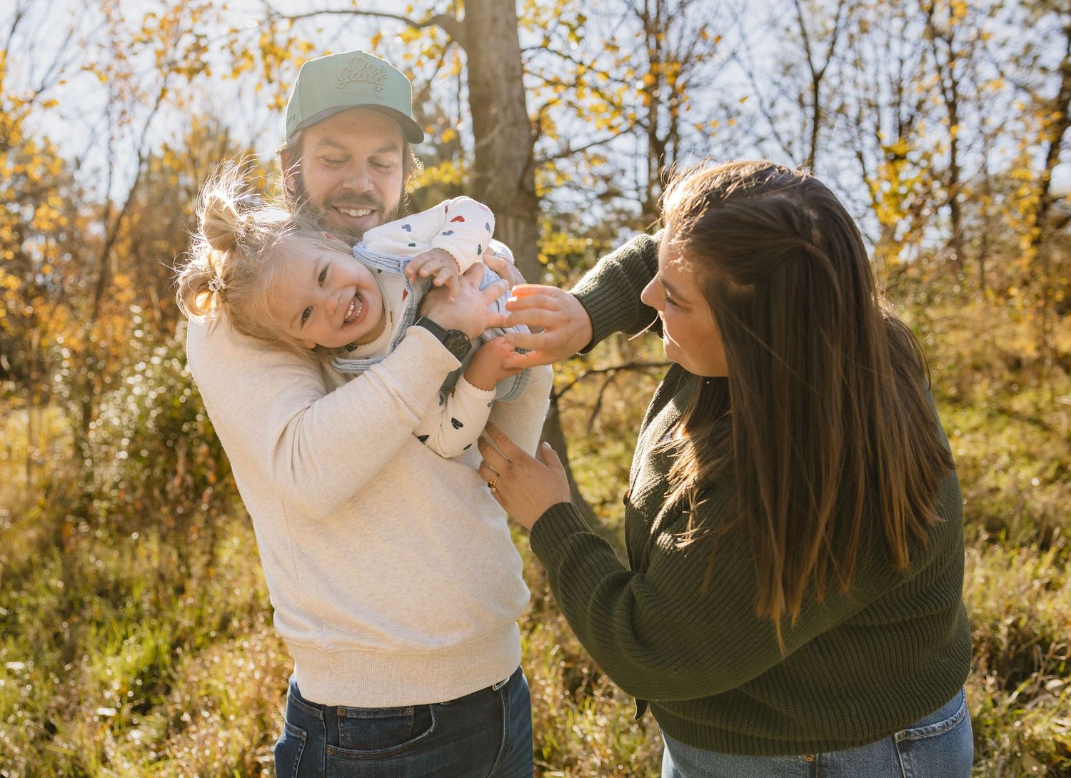 A joyful family with a laughing toddler being playfully held by parents outdoors in a sunny autumn setting.