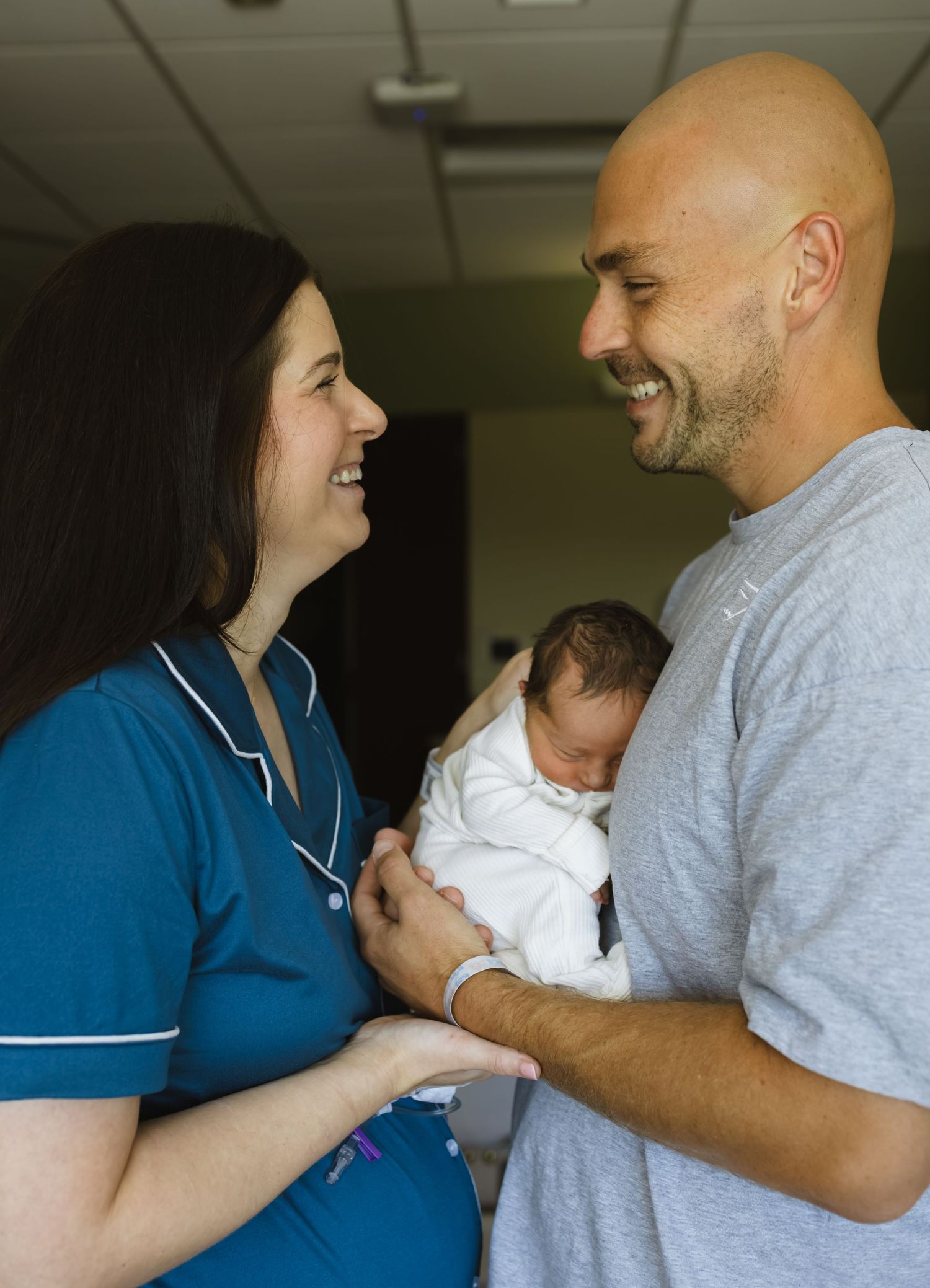 A nurse in blue scrubs smiles at a bald man holding a newborn baby in a hospital room.