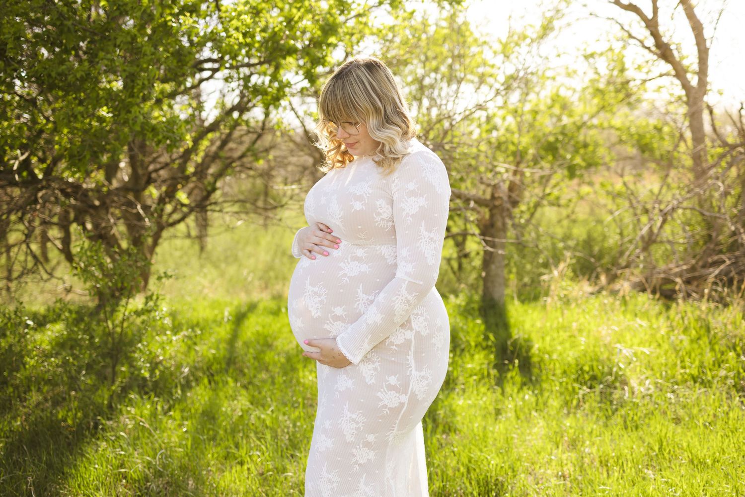 Pregnant woman in white lace gown cradling baby bump outdoors in sunlit green field during maternity photo session.