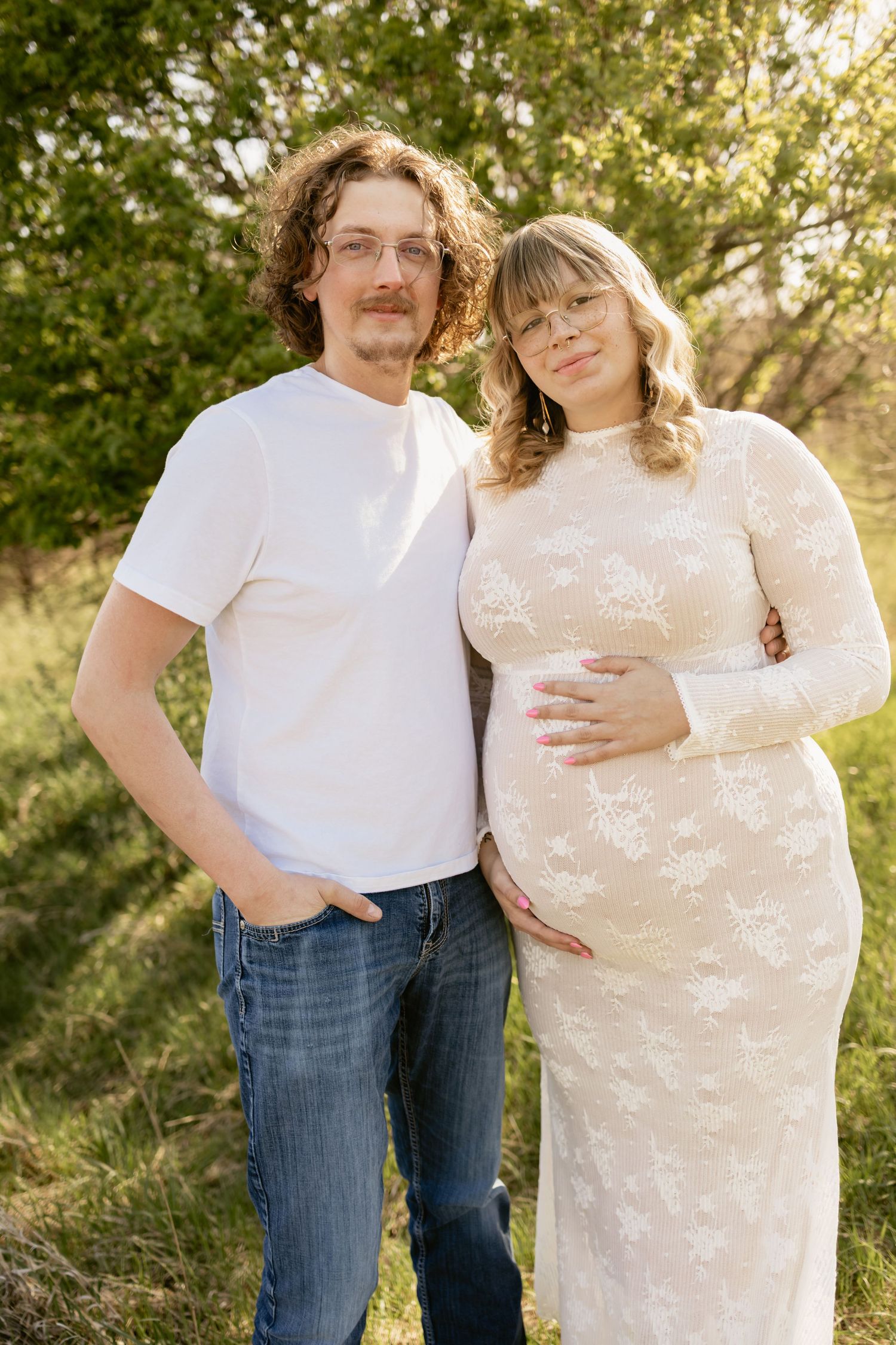 A pregnant couple poses outdoors, woman in white lace dress cradling belly, man in white tee and jeans, surrounded by greenery.