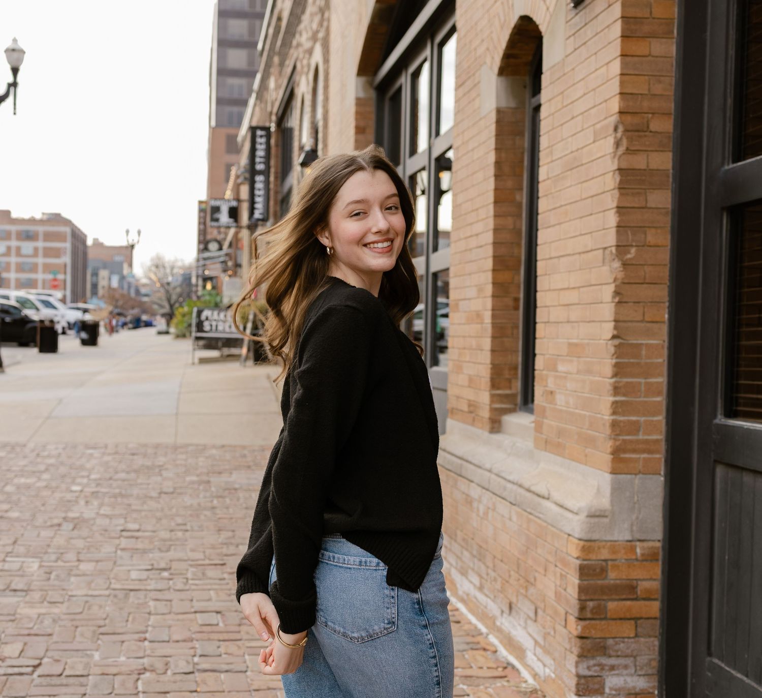 Young woman in black sweater and jeans smiling on a brick sidewalk outside a historic building.