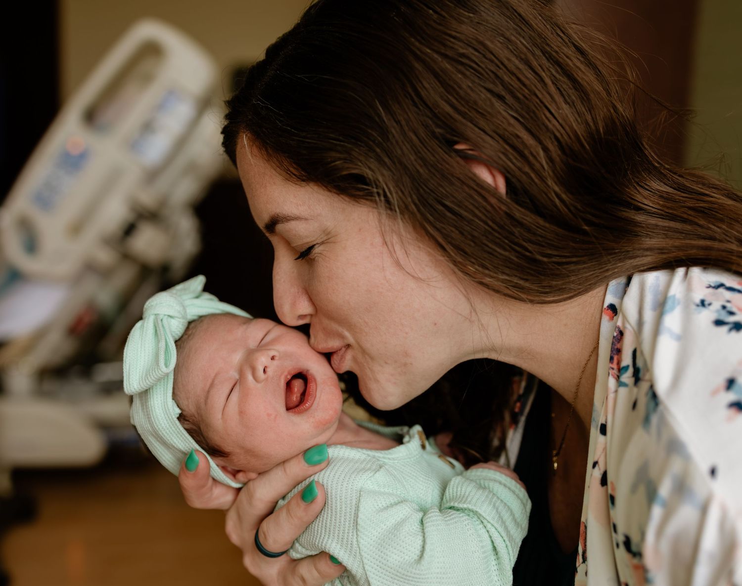 New mother kissing her yawning newborn baby wearing a mint green bow in a hospital room for their Fresh 48 photos in Omaha, NE.
