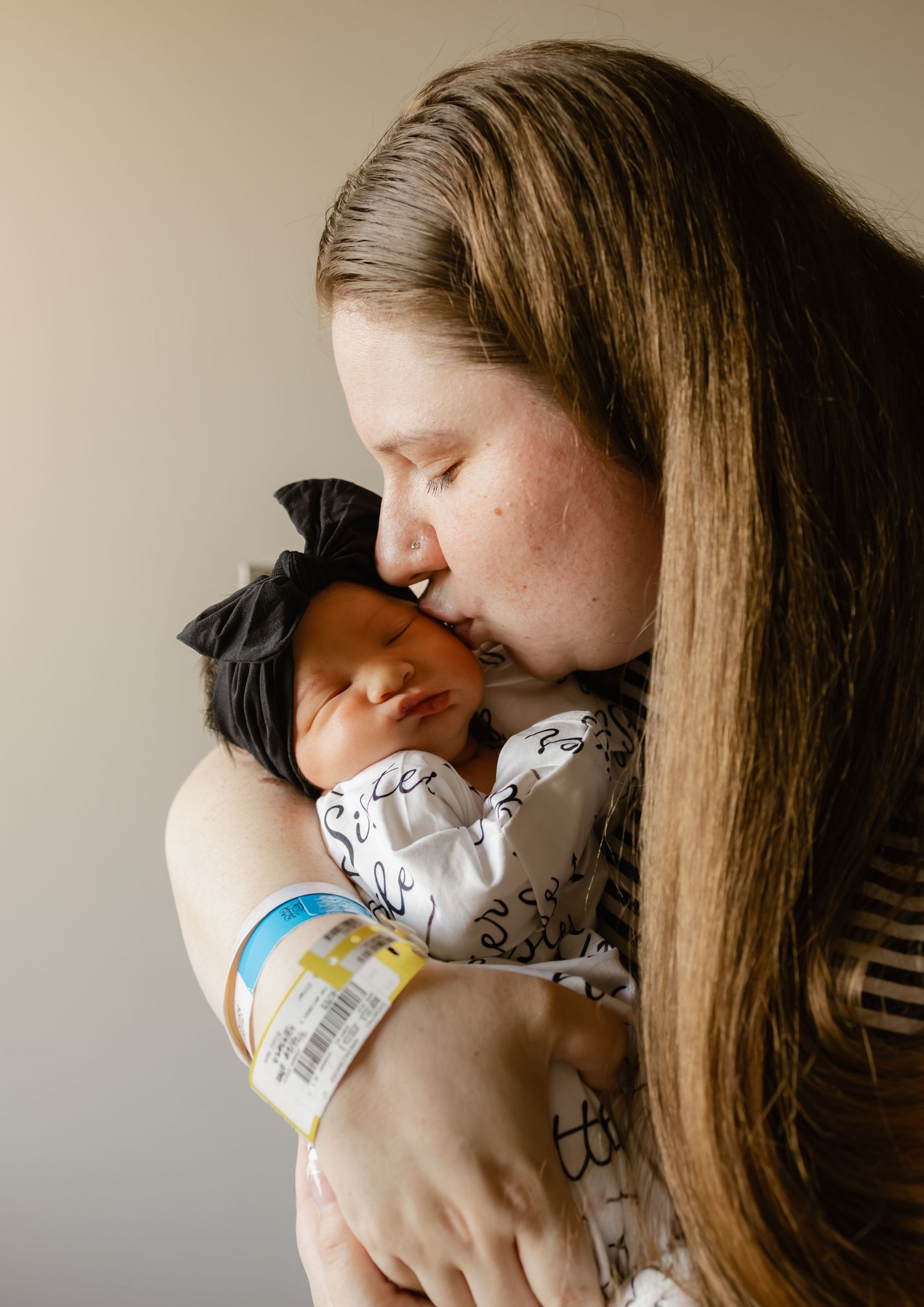 A new mother with long hair gently kisses her sleeping newborn baby dressed in white, in a tender hospital room moment.