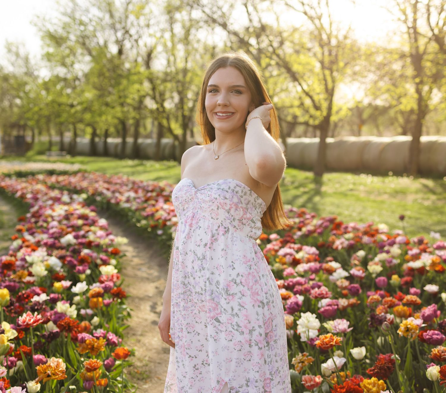 Young woman in floral dress smiling among colorful tulip rows in sunlit garden.