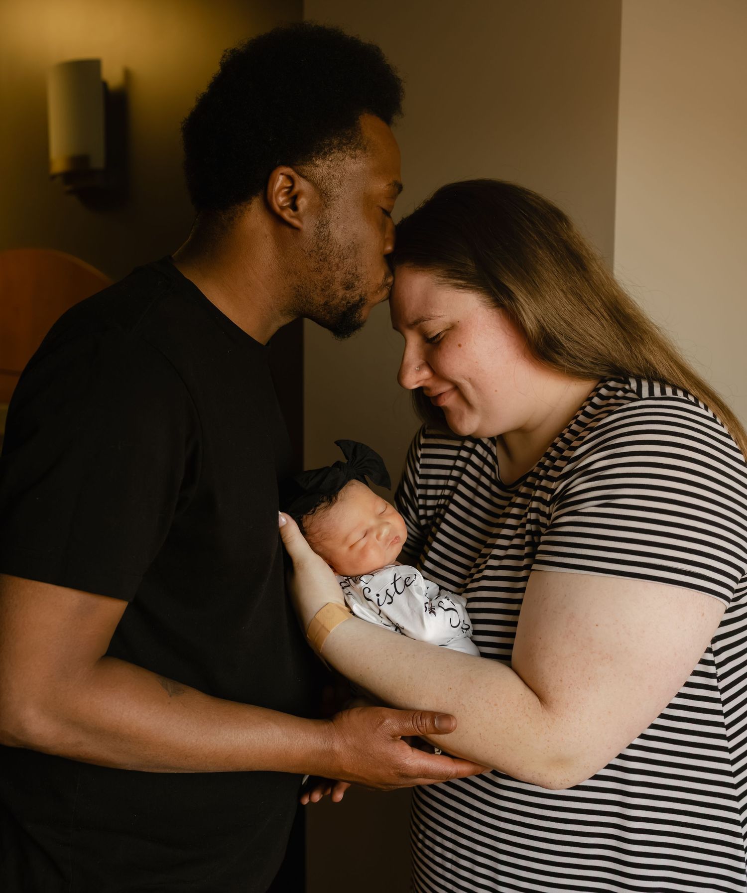 A couple tenderly holds their newborn baby, with the father kissing the mother's forehead in a warm indoor setting.