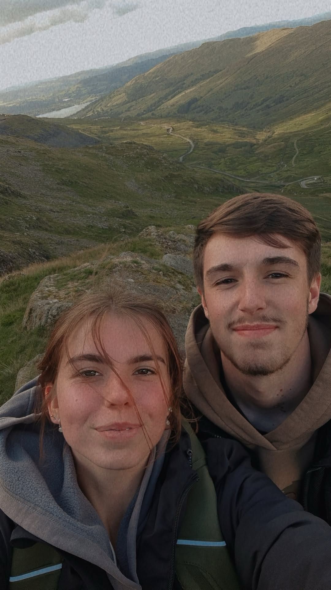 Two young people smile for a selfie with a scenic mountain valley landscape behind them.