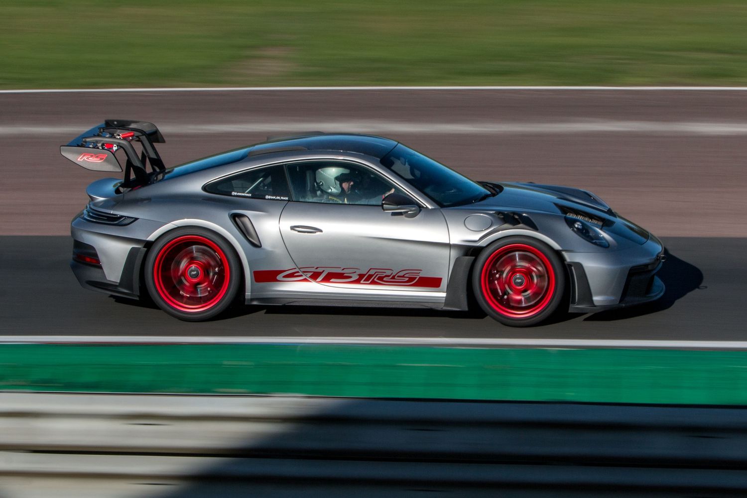 Silver Porsche 911 GT3 RS with red wheels and racing livery speeding along a motorsport circuit track.