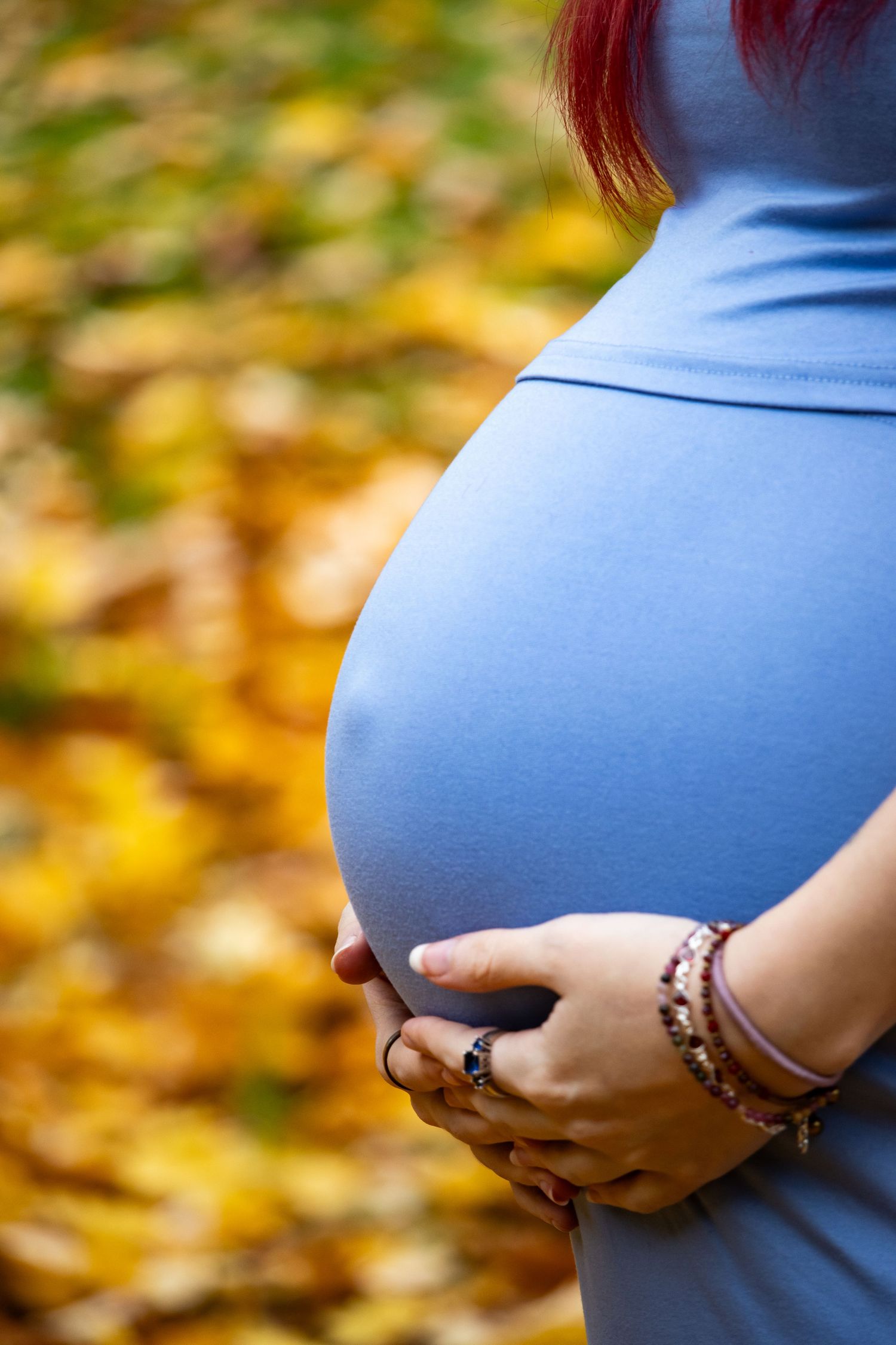 Pregnant woman in blue dress cradling belly against autumn leaves background.