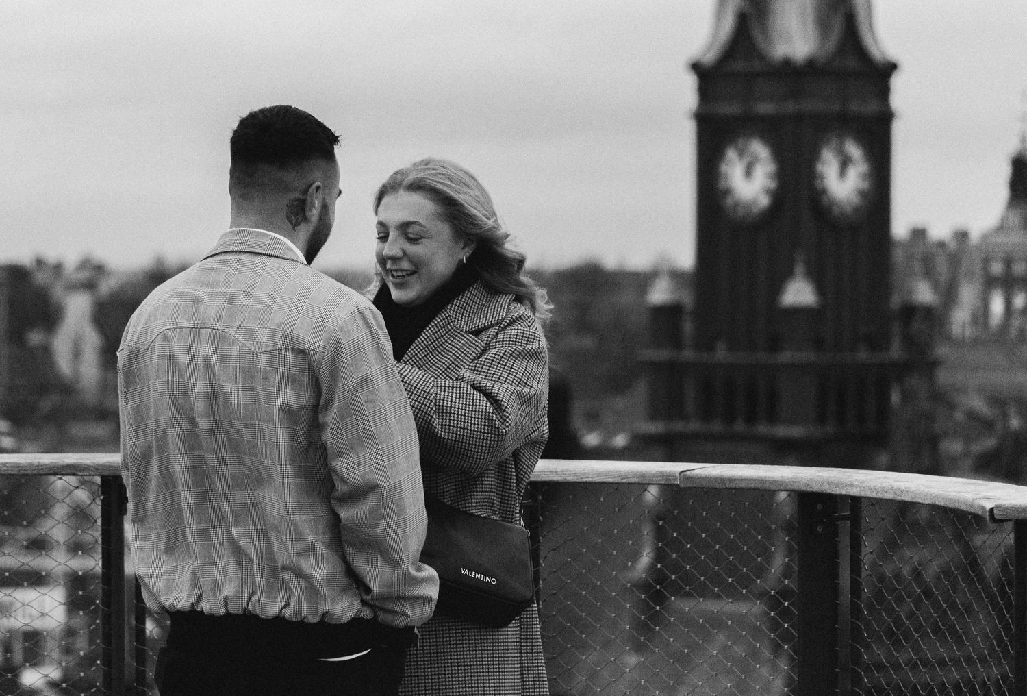 Black and white photo of a couple sharing a romantic moment on a rooftop with a clock tower in the background.