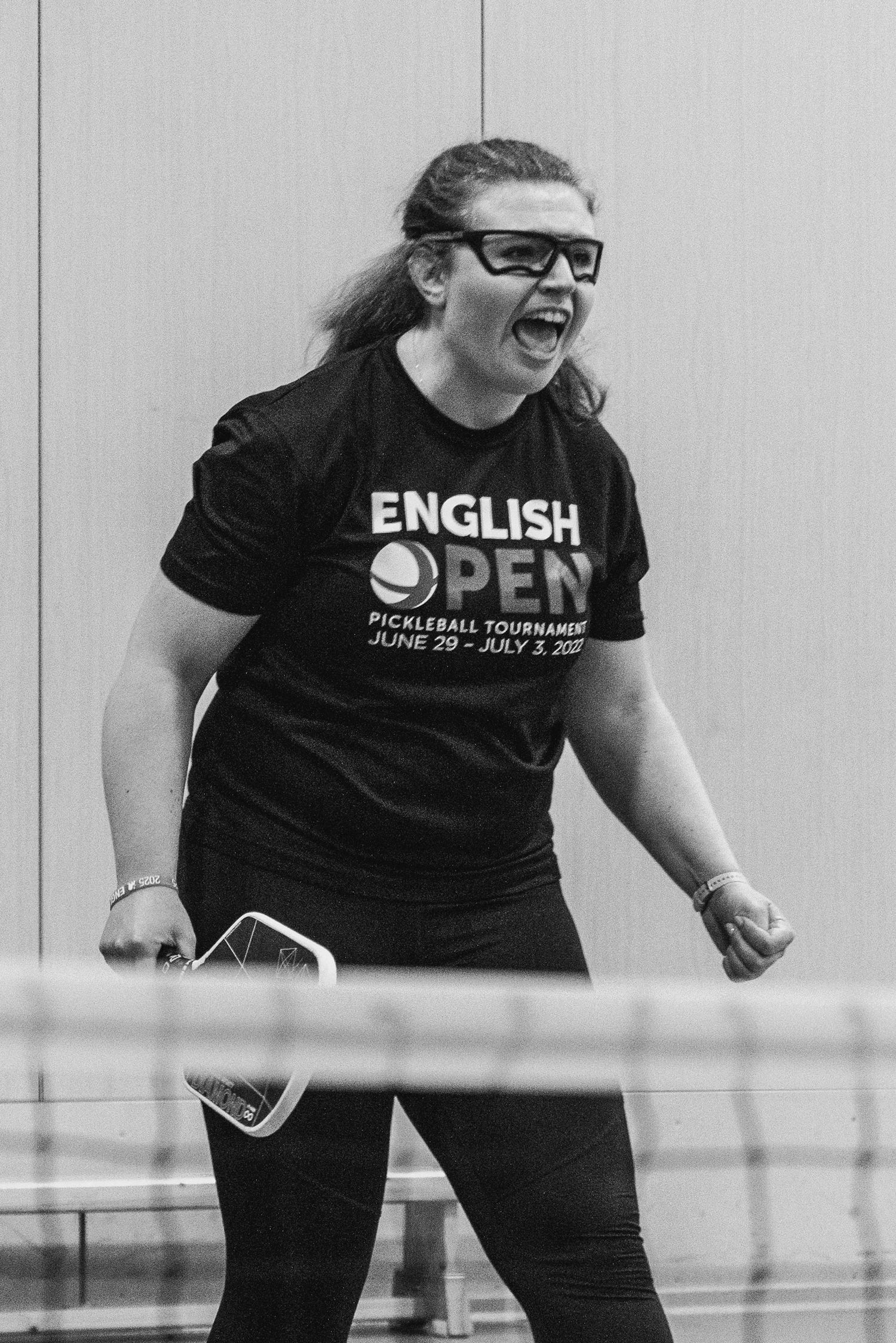 Woman celebrating at English Open Pickleball Tournament, wearing black t-shirt, glasses, holding paddle near net.