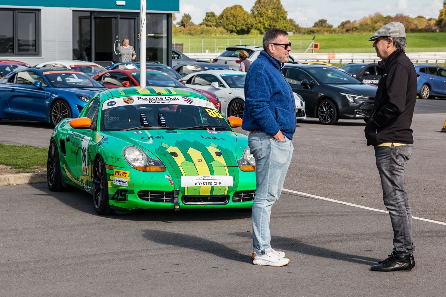 Two men standing beside a green Porsche Cayman race car in a paddock area.