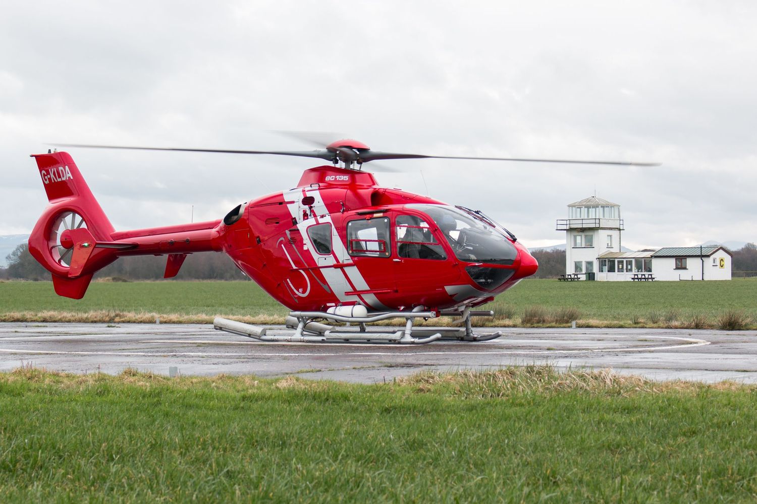 Bright red emergency helicopter parked on airfield with buildings in background.