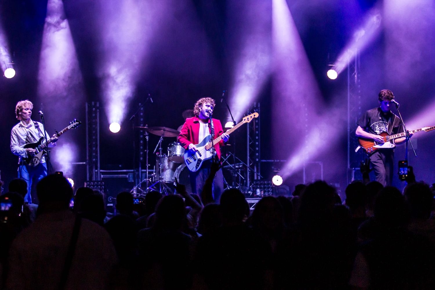 Rock band performing on stage with dramatic purple lighting and silhouetted crowd.